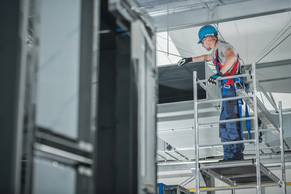 Commercial HVAC technician servicing ductwork from a scaffold inside a large industrial facility.