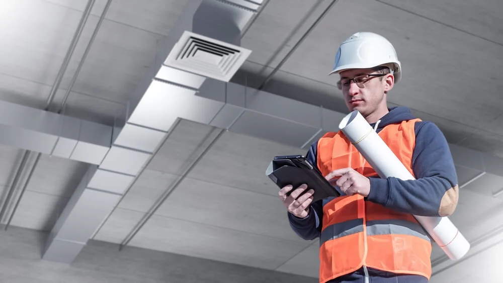 Commercial HVAC technician reviewing ductwork installation plans on-site, standing under exposed ceiling ventilation system.
